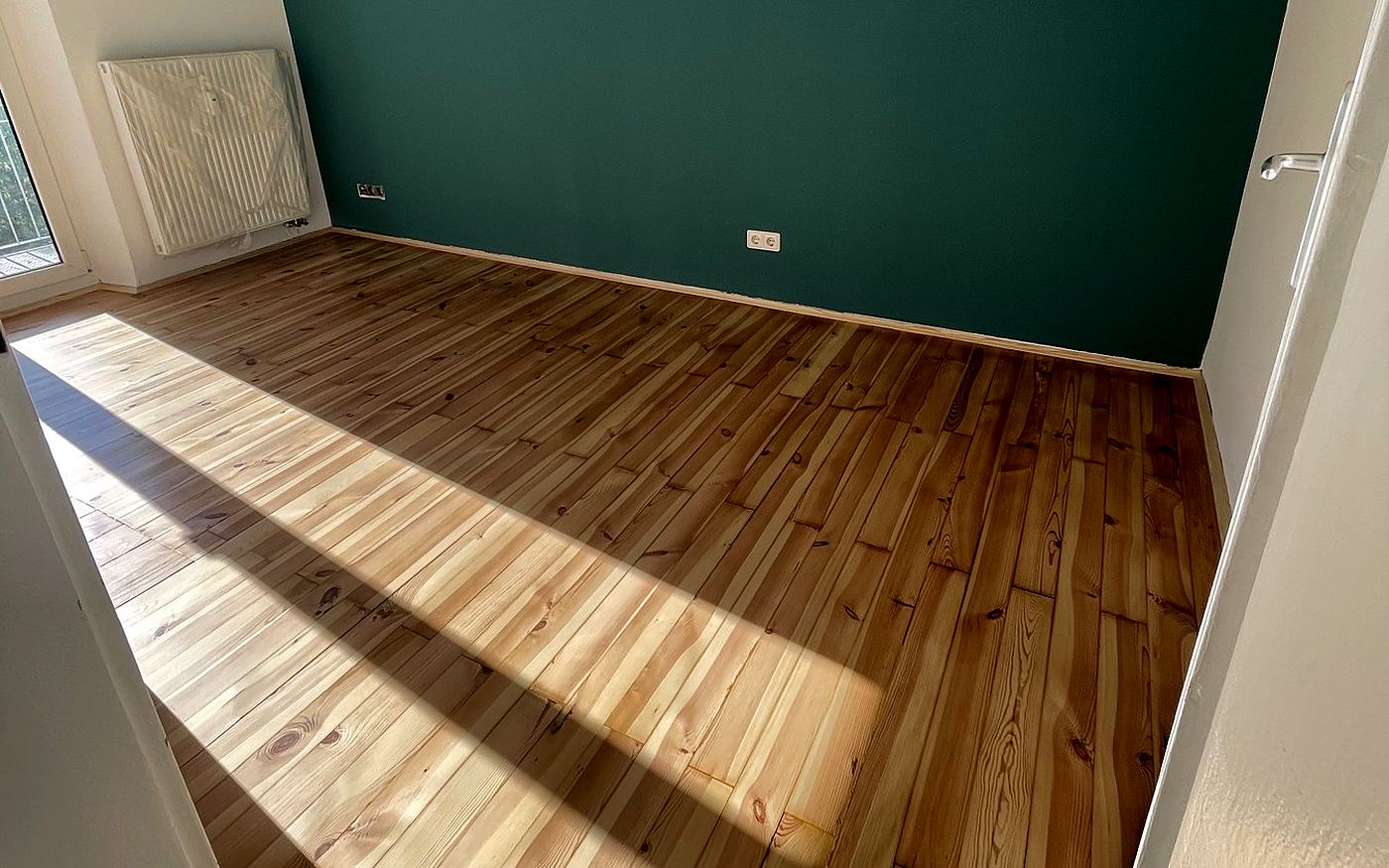 Frisch versiegelter heller Dielenboden mit Sonnenstreifen vor grüner Wand